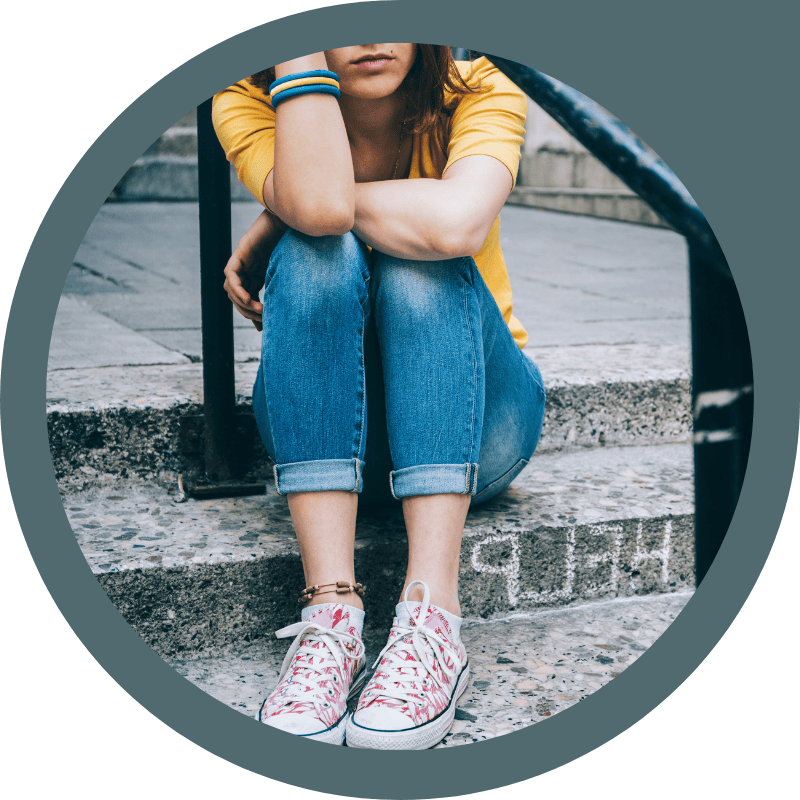 A person wearing a yellow shirt, blue jeans, and patterned sneakers sits on concrete steps with arms resting on knees, face partially hidden.